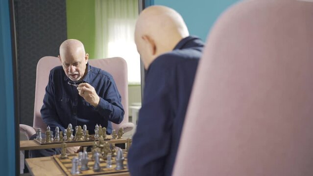 Focused Old Man Playing Chess Alone At Home And Looking At Himself In Mirror.
The Old Man Is Playing Chess By Himself In Front Of The Mirror.
