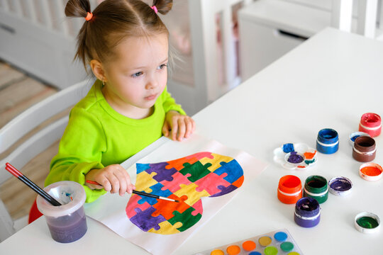 The child draws a picture with a thoughtful look looking ahead. A girl paints a heart out of puzzles with a brush and watercolor paints. Drawing is a symbol of autism