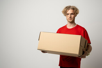 Portrait of a young delivery man, courier in red cap and t-shirt standing with parcel post box isolated over white background.