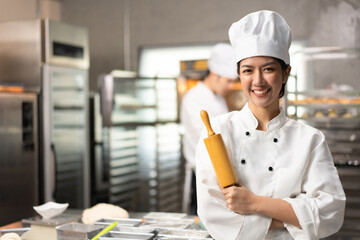 Selective focus of Asian female baker in white chef uniform, standing with arms crossed holding a rolling pin and smiling at camera, with blurred bakery kitchen in background. Copy space on left side.