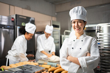 Selective focus of Asian female baker in white chef dress and hat, standing with arms folded and smiling at camera, with blurred colleagues kneading dough in the background. Copy space on left side.