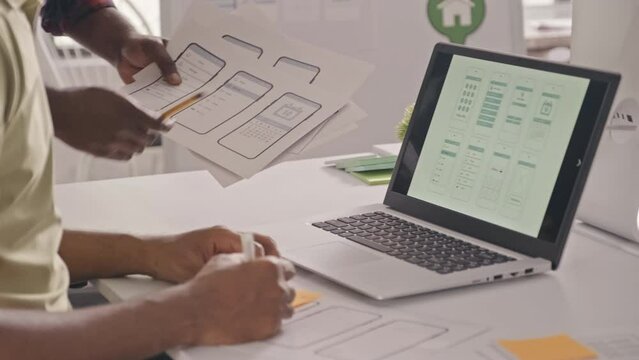 Cropped shot of two male UX designer discussing scheme of mobile app interface on paper and laptop and drawing wireframe while working together on IT project in office