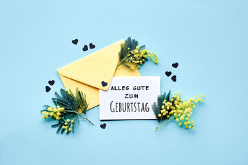 Text Alles Gute zum Geburtstag means Happy Birthday in German language. Text Special moments on paper card with mimosa flowers in yellow envelope on mint blue background, confetti. Flat lay, top view.