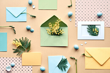 Easter background with quail eggs. Mimosa and freesia flowers, mint green envelopes and color paper on yellow background. Flat lay, overhead, knolling, neatly arranged springtime decor.