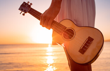 Young female girl holding guitar ukulele on the beach looking out to the sunset. Relax on a summer ocean vacation concept.  - obrazy, fototapety, plakaty