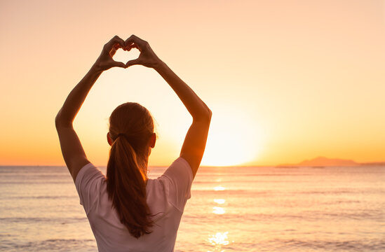 Young woman on the beach making heart shape facing the golden sunset. I love nature and life. 