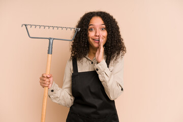 Young african american gardener woman holding a rake isolated is saying a secret hot braking news and looking aside