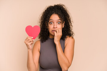 Young african american woman holding a heart for valentines day isolated biting fingernails,...