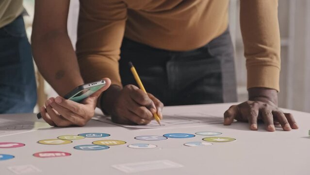 Cropped shot of team of mobile app developers holding smartphone and making notes on sketches while working on UX design project at office table