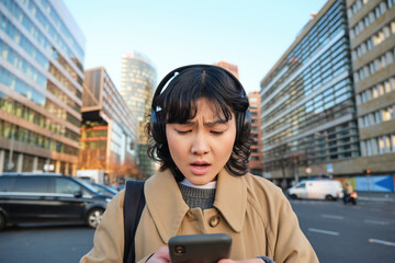 Portrait of young asian girl, student walks in city, listens music in headphones and uses mobile...