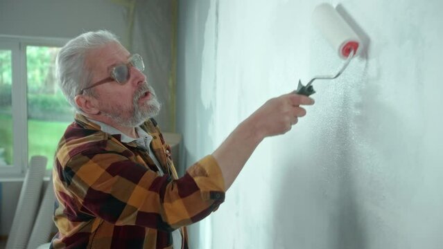 Elderly man is painting wall with white paint using paint roller. Male pensioner is making repairs to his apartment against the backdrop of a window with bright sunlight. Side view.