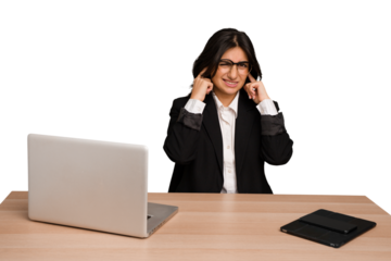 Young indian woman in a table with a laptop and tablet isolated covering ears with fingers, stressed and desperate by a loudly ambient.