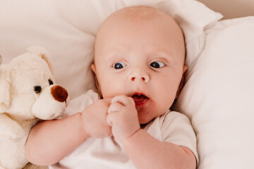 Little boy funny cute child baby lying on bed pillows with fluffy toy. Playful toddler with bulging eyes having fun, making faces grimaces. Happy childhood, family concept
