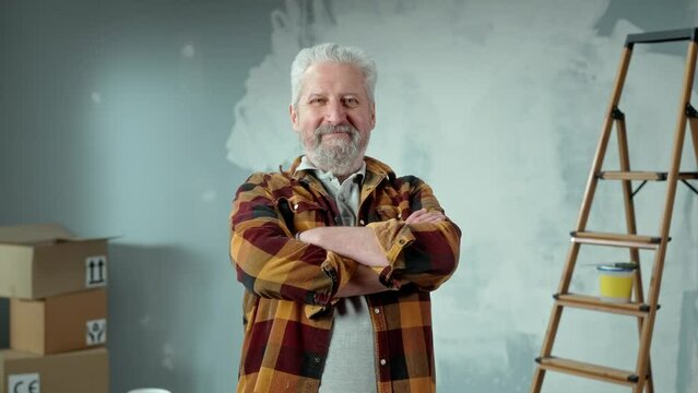 Elderly gray haired man with beard looking at camera, crossing arms over his chest and smiling. Portrait of pensioner posing in an apartment against background of wall painted with white paint.