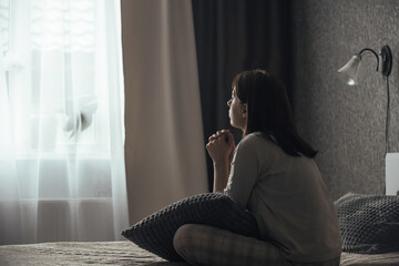 Naklejka premium A sad young woman in pajamas with seasonal affective disorder or depression sits alone on the bed and looks out the window. The concept of winter depression due to lack of sunlight