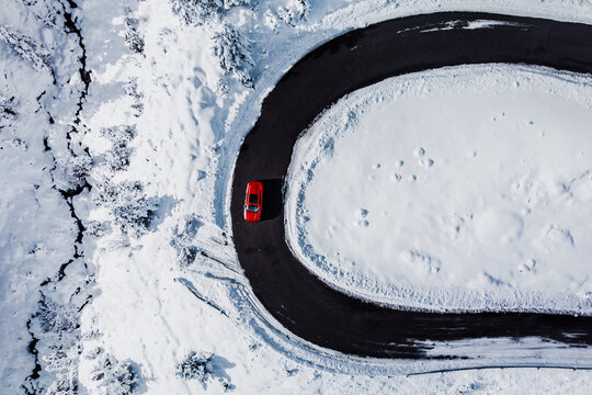 Car in the curve of a snowy winding road in a winter landscape of a mountain pass. Top down aerial view of a red passenger car in a snow. Vacations, travel, drive, route, journey and tourism concept.