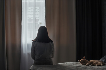 A sad young woman with seasonal affective disorder or depression sits alone on the bed and looks out the window, a domestic cat lies nearby. The concept of winter depression due to lack of sunlight