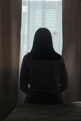 Silhouette of a young woman in pajamas against the background of a window at home on a bed with...
