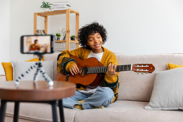 Blogger guitarist. African american girl blogger playing guitar talking to webcam recording vlog. Social media influencer woman streaming at home indoors. Music content creator broadcast tutorial.