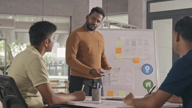 Indian UX designer standing by flipchart in office and giving presentation to team during business meeting