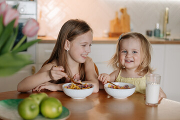 two sisters in pajamas having breakfast in the kitchen eating colorful cereal with milk at home in...