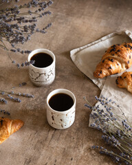 Two cups of black coffee with chocolate croissant on brown wood background with composition of lilac lavender flowers