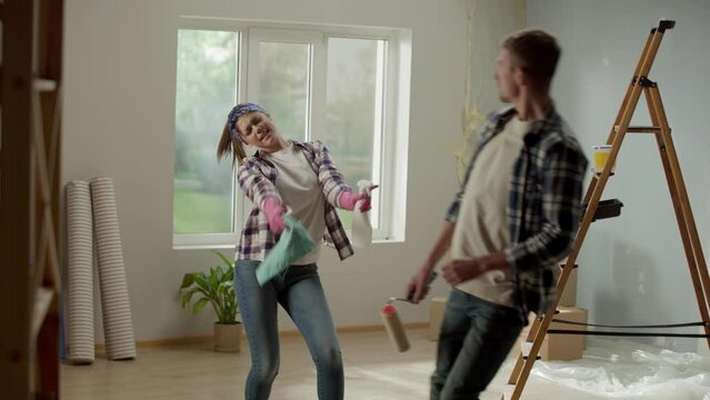 Young woman with rag and spray bottle is singing and dancing merrily. Man joyfully is singing into paint roller as if into microphone. Couple are having fun cleaning up an apartment during renovations
