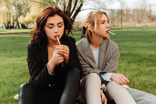 Two pretty women friends sitting on grass smoking cigarette. Couple of gay lesbian girls hugging embracing together girlfriends, have a date. LGBT concept. Addiction, cancer, tobacco. Bad habit
