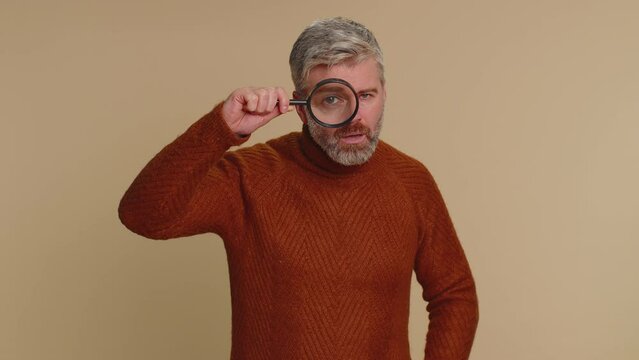 Investigator researcher scientist old man holding magnifying glass near face, looking into camera with big zoomed funny eyes, searching, analysing. Senior middle-aged guy on beige studio background