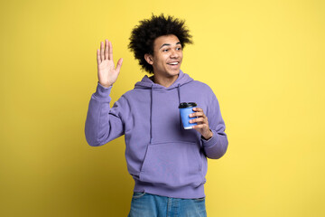Smiling African American man wearing stylish hoodie, holding cup of coffee waving hand, isolated on yellow background