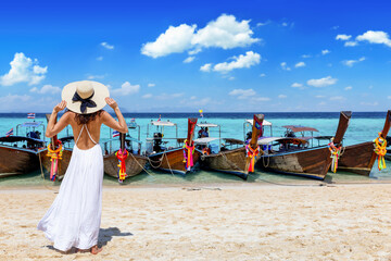 Thailand travel concept with a woman in a white summer dress standing on a beautiful beach in front of longtail boats waiting for tourists