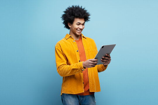 Smiling African American man holding digital tablet ordering food, shopping online isolated on blue background. Happy modern hipster using mobile app, watching videos, rearing e-book. Technology   