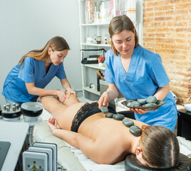 Two masseuses working at the spa salon massage the legs and perform a stone therapy procedure a...