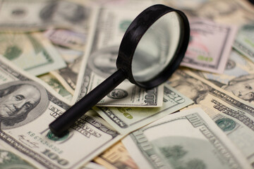 Dollars lie in a large pile forming a solid background. On dollar bills, there is a magnifying...
