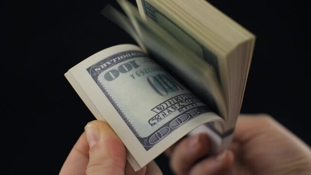 On a black background, a hand counts while flipping through a stack of one hundred dollar bills. A stack of dollars in hand is flipped through in slow motion with a finger. Dollar bill concept.