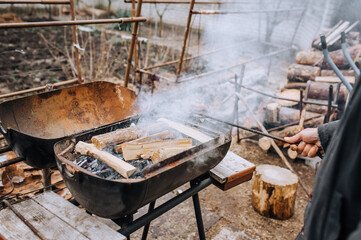 A man in the open air, in nature, holds a metal poker in his hands and corrects burning firewood in the barbecue. Photography, food preparation.