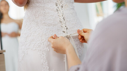 Friends helping woman with wedding dress knot in a room or boutique for marriage ready, fitting or...