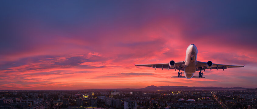 Airplane is flying in colorful sky over the city at night. Landscape with passenger airplane, skyline, purple sky with red and pink clouds. Aircraft is landing at sunset. Aerial view. Transport