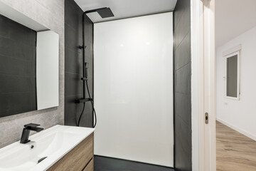 Newly renovated bathroom with black faucets, square mirror, gray and black walls and floors, glass partition, and light wood cabinets with drawers