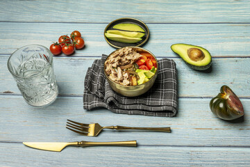 A delicious canned tuna and tomato salad in a golden bowl with matching cutlery on a black cloth on the blue wooden table with tomatoes, half an avocado and a glass of water