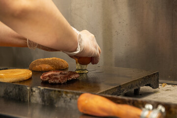 Hands of a chef flipping beef burgers on a restaurant griddle and dropping an egg to fry