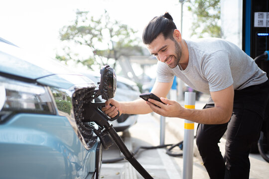 Handsome Asian young man holding an EV plug connector and attaching onto the car, man plugged in the EV charging connector to the vehicle.
