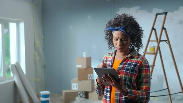 African American woman is using tablet to plan renovations in apartment. Black female in checkered shirt thinking, viewing and visualizing ideas against background of apartment in process of