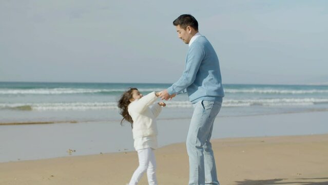 Asian dad and cute little girl having fun at seashore on sunny day. Bearded man twirling young child under his arm while daughter laughing and enjoying time with loving father. Parenthood concept