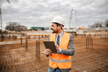 Obraz na płótnie Canvas A happy engineer is checking building works on tablet while standing on construction site.