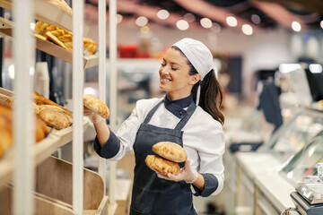 A baker is filling up the shelves with bread while standing at bakery department in supermarket.