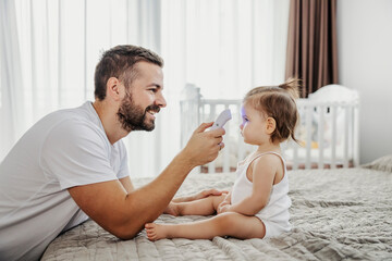 A happy father is measuring temperature with digital thermometer while toddler is sitting on a bed.