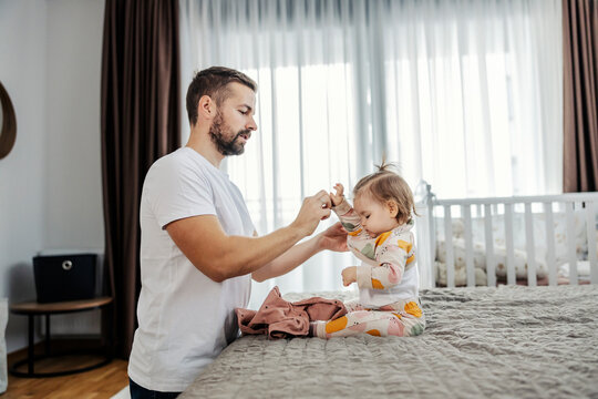 A father is changing the little girl's clothes while she is sitting on a bed.
