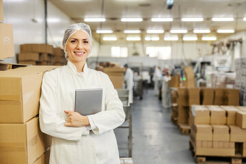 A food factory supervisor is leaning on boxes with goods while holding tablet and smiling at the...