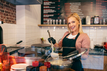 A happy pancake shop worker is holding ladle with crepes mixture and smiling at the camera.